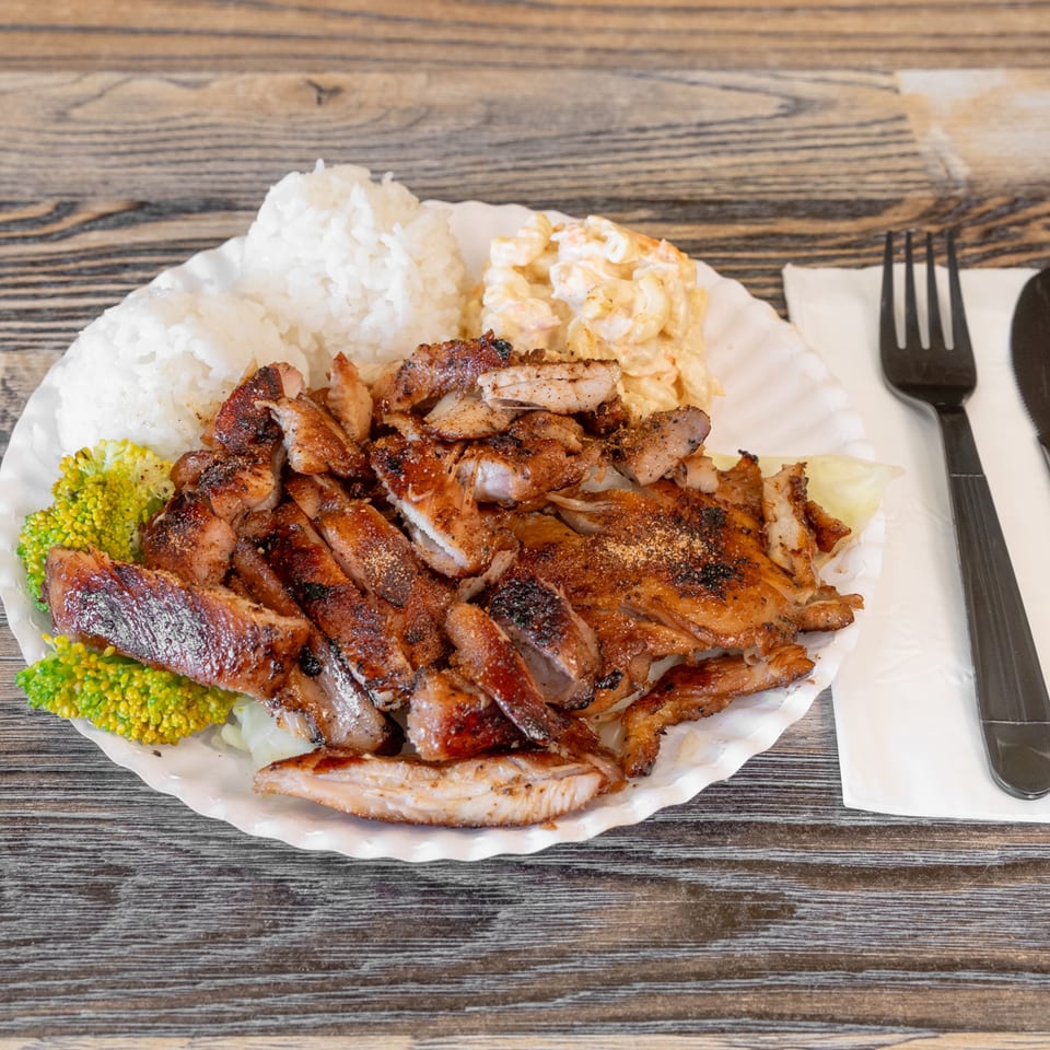 Plate of sliced grilled meats with white rice, broccoli, creamy salad, and utensils on a rustic wooden table.