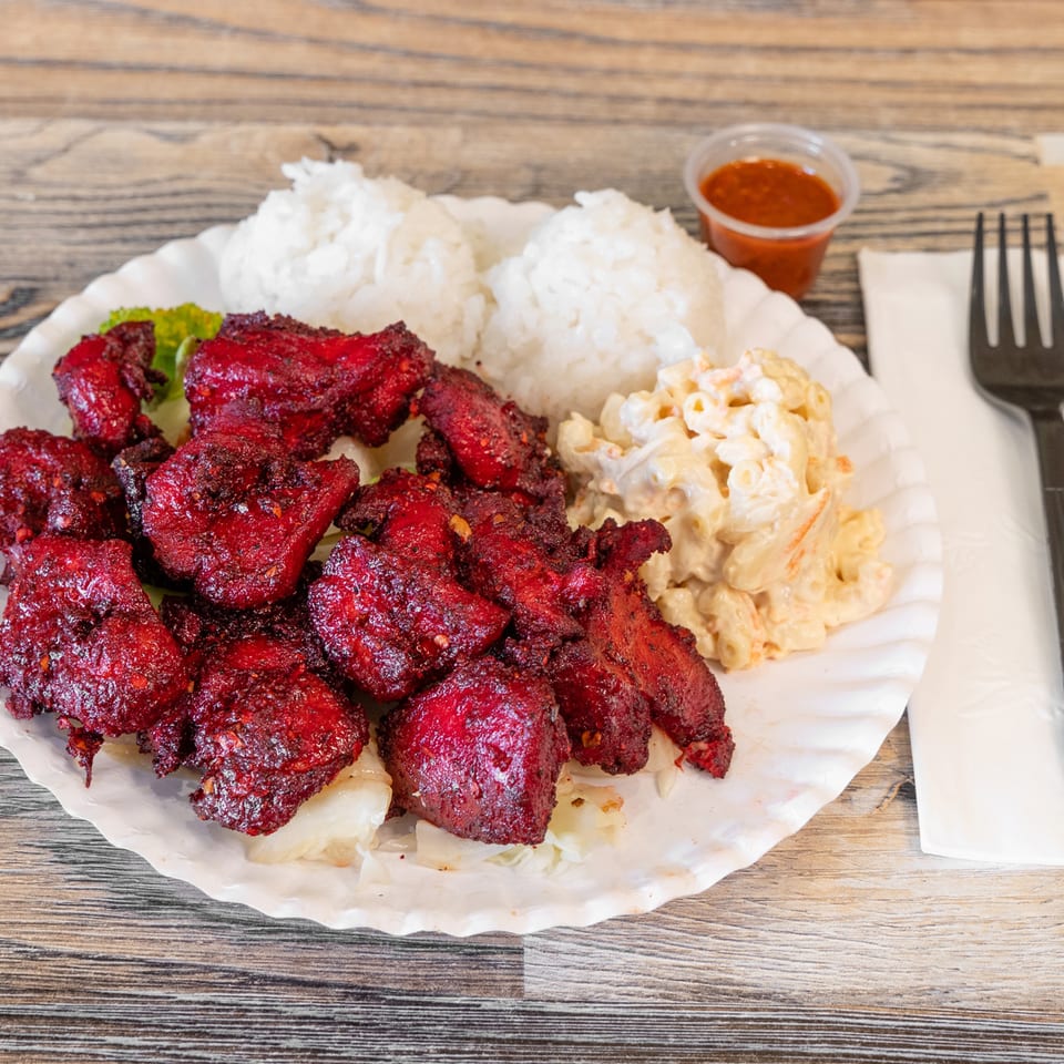 Plate of red-spiced grilled chicken with white rice, macaroni salad, lettuce, and dipping sauce on a wooden table.