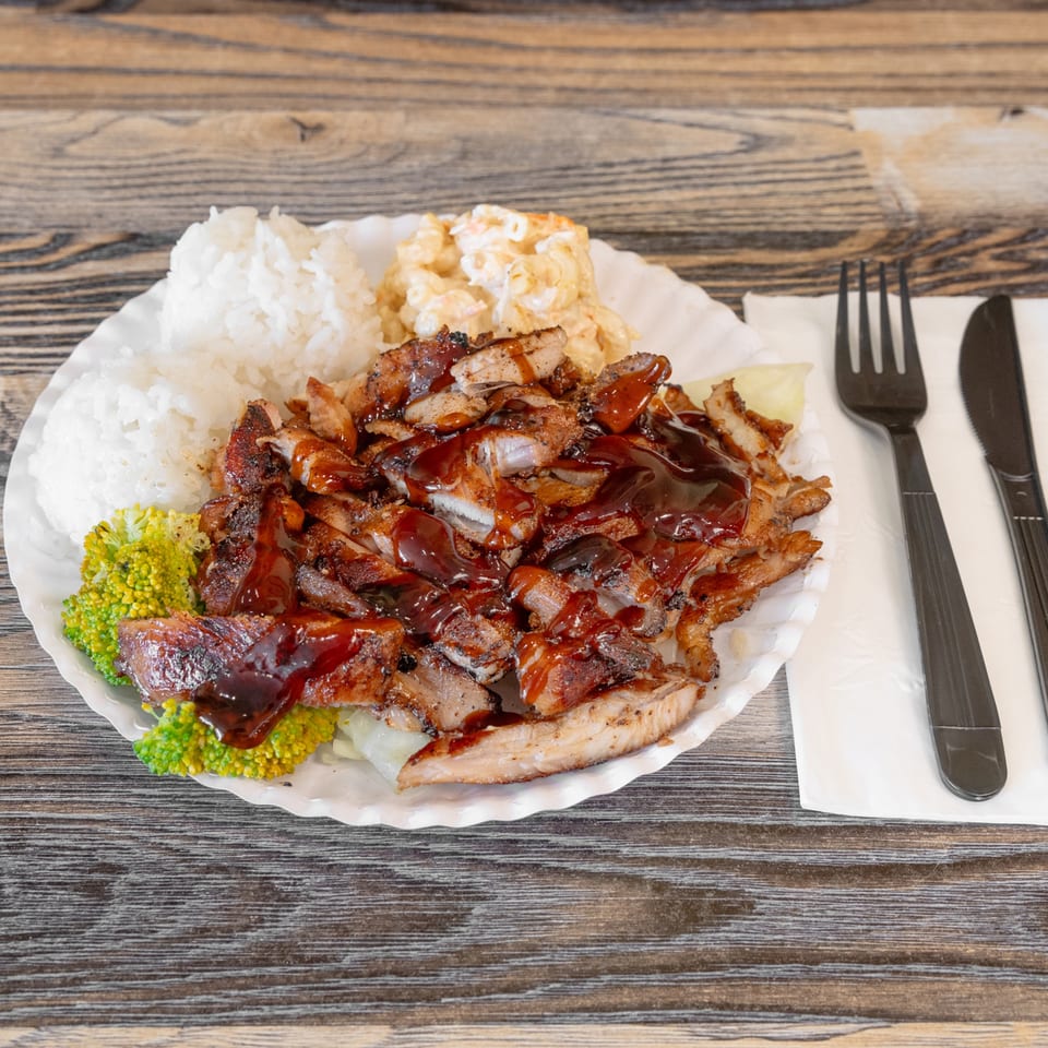 Plate of barbecue chicken and pork with rice, macaroni salad, broccoli, and sauce on a wooden table.