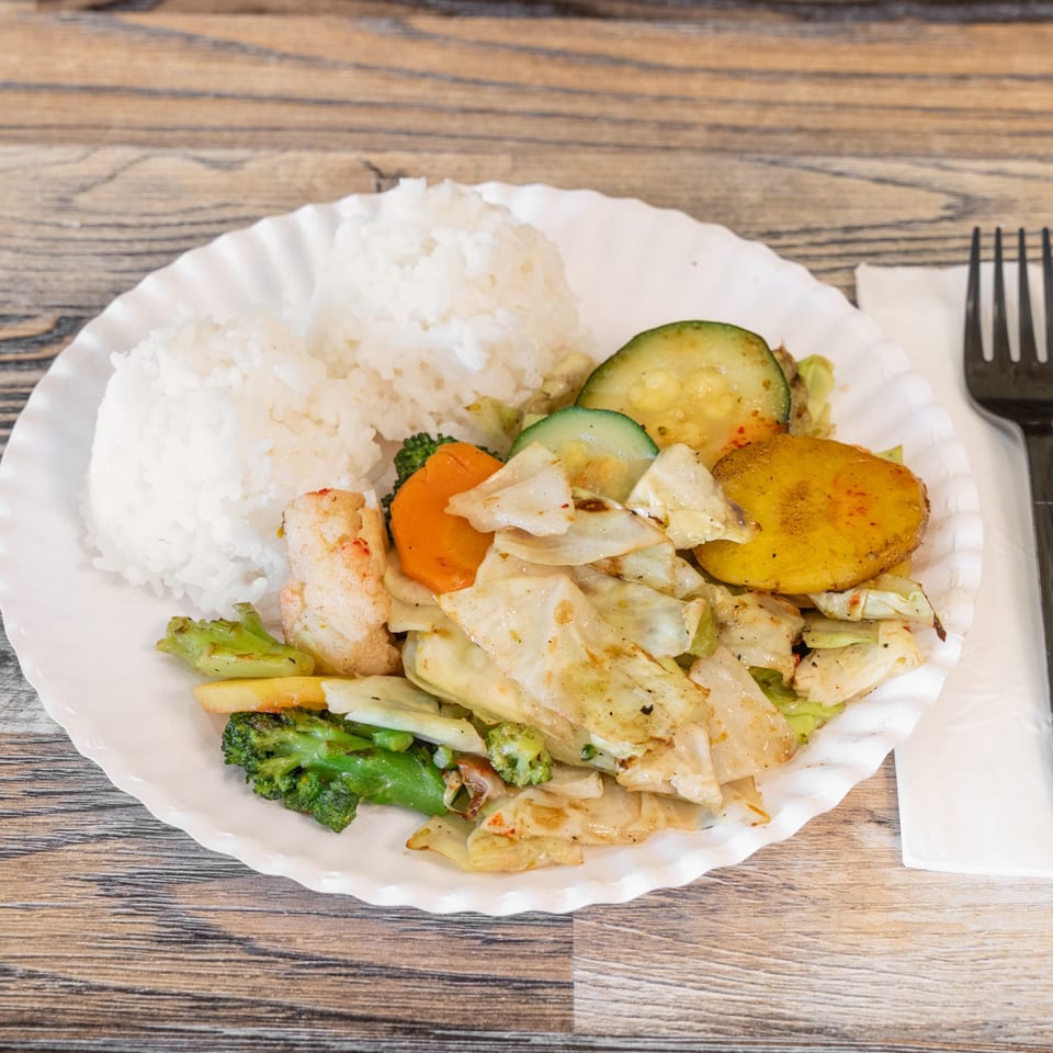 Plate of white rice, sautéed shrimp, mixed vegetables, and roasted potato on a wooden table with fork and knife.