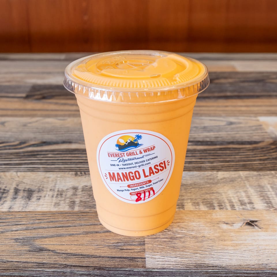 Mango lassi in a clear takeout cup labeled Everest Grill & Wrap, resting on a rustic wooden table.