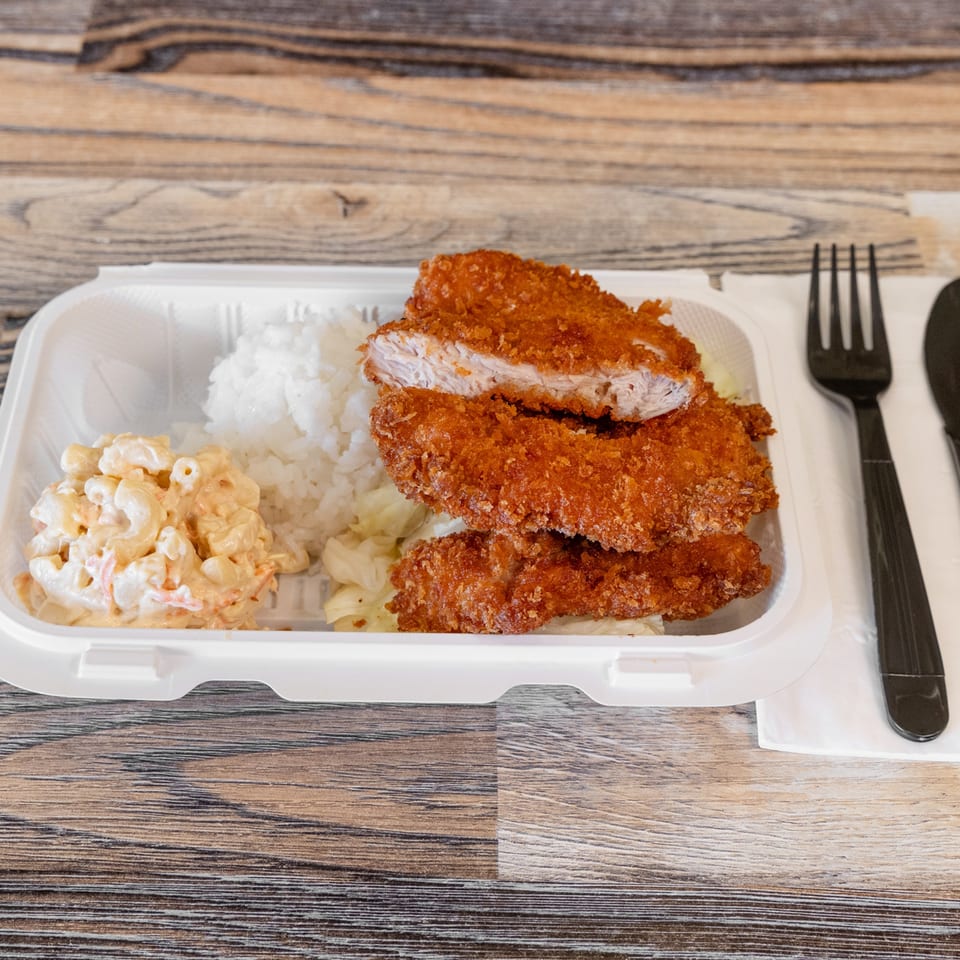 Takeout container holding breaded fried chicken, white rice, macaroni salad, and cabbage, with black utensils on a wooden table.